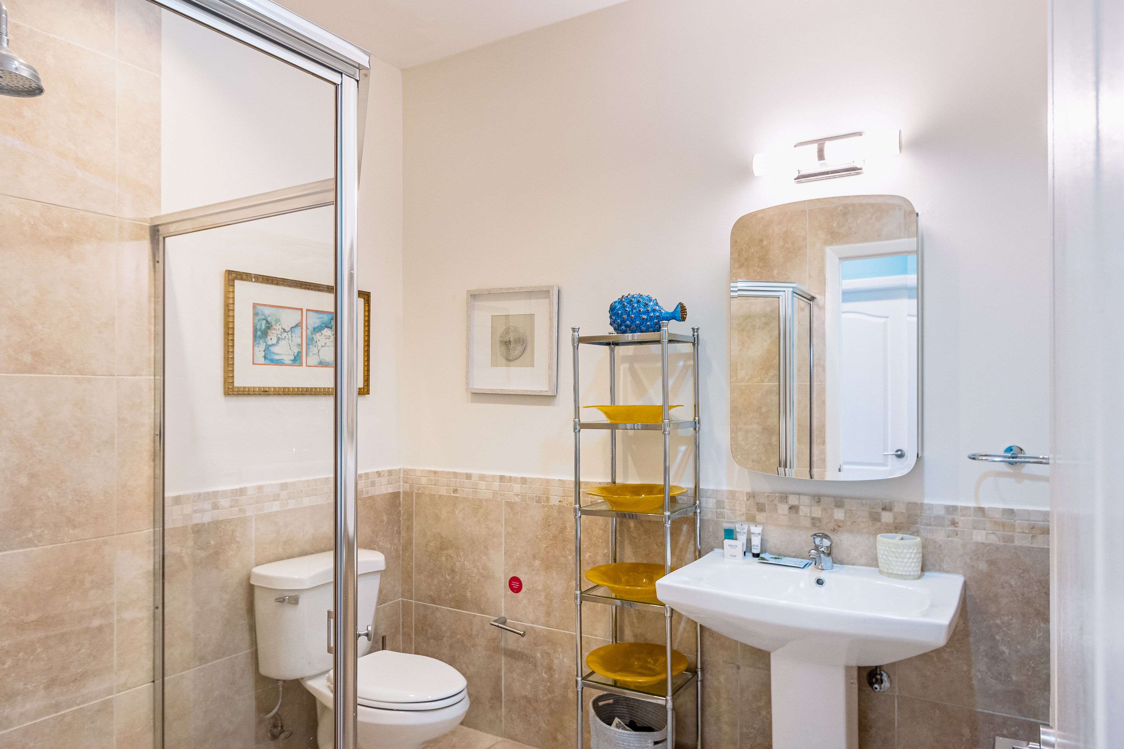 A bathroom with a glass shower enclosure, a toilet, a sink with a mirror, and shelves with yellow bowls.