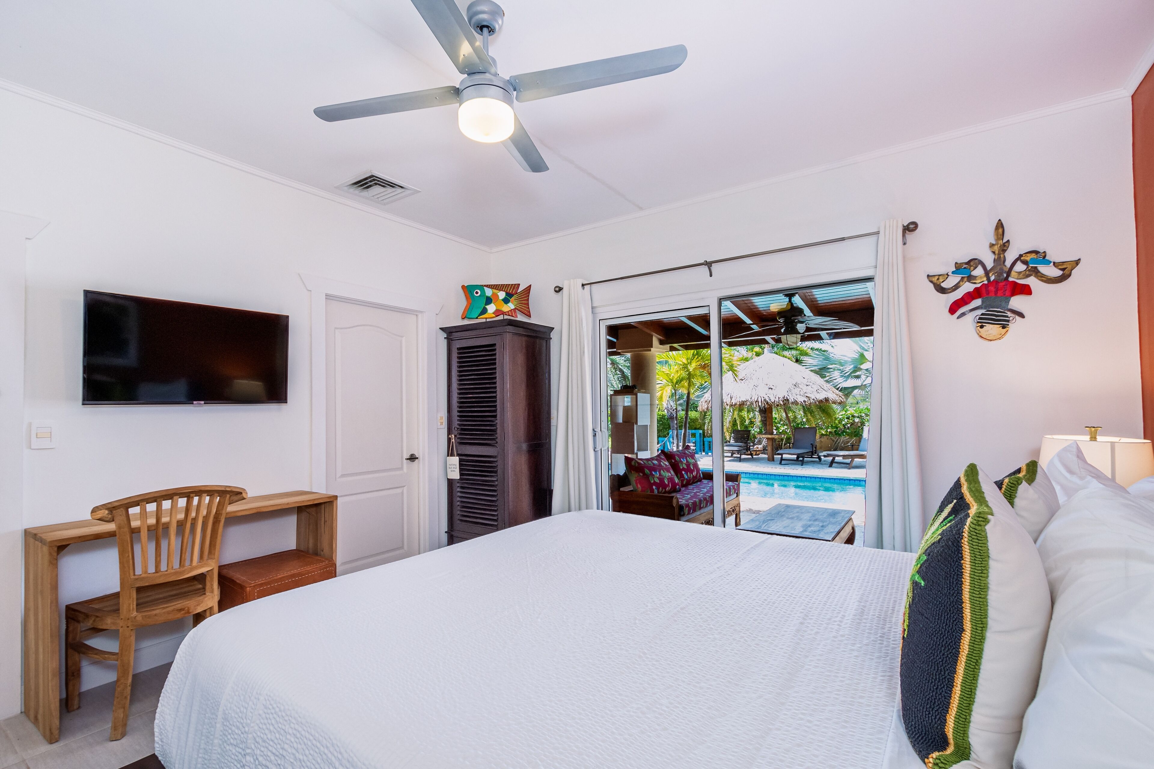 A bedroom with a bed, a desk, a chair, a TV, a ceiling fan, and a view of an outdoor pool area.