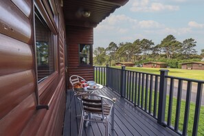 Una cabaña de madera con balcón, una silla de metal, una mesa con cosas para el desayuno y vistas del paisaje circundante.
