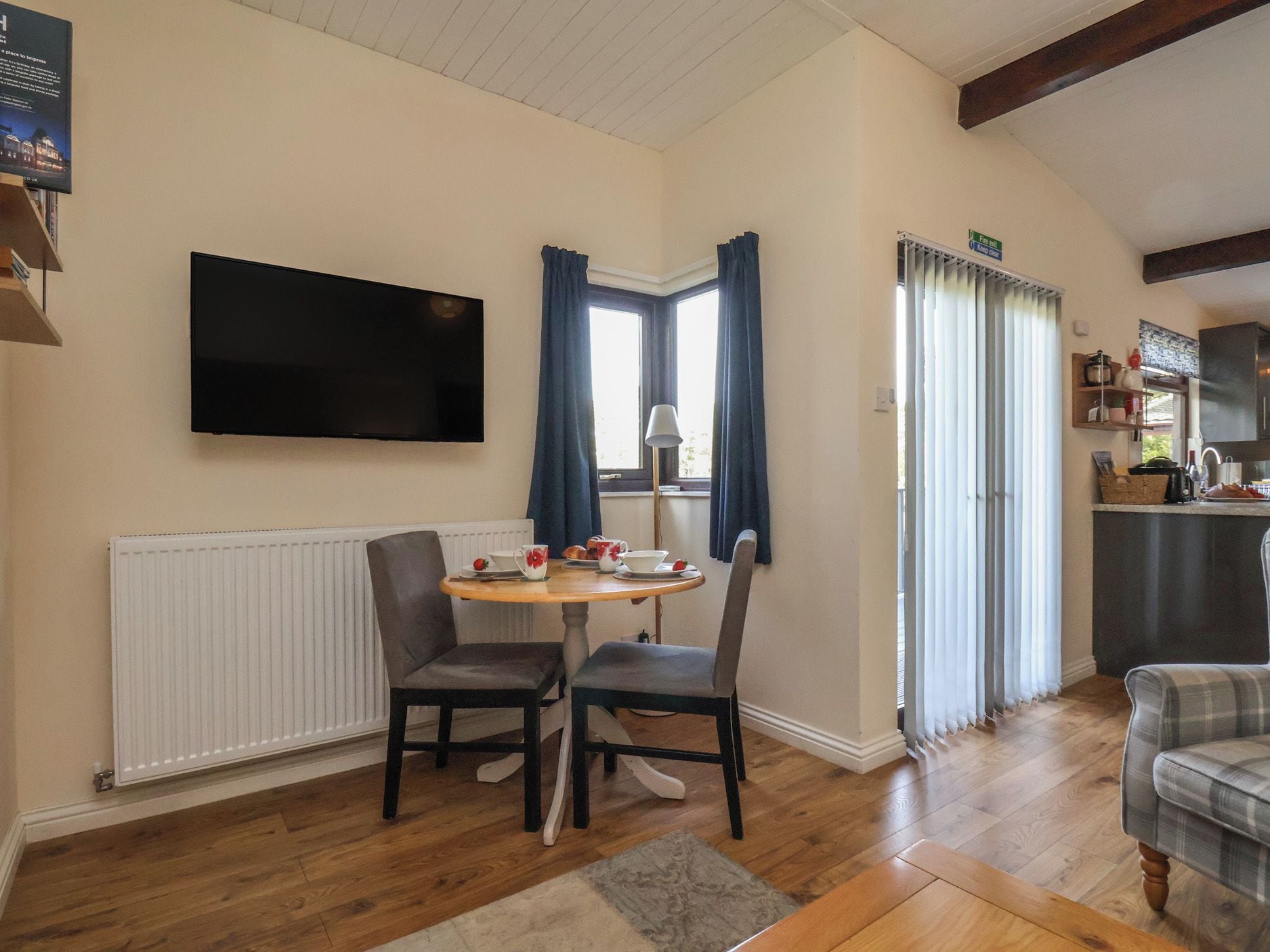A room with a dining table, two chairs, a flat-screen TV mounted on the wall, a window with curtains, and a kitchen area with cabinets and a countertop.