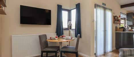A room with a dining table, two chairs, a flat-screen TV mounted on the wall, a window with curtains, and a kitchen area with cabinets and a countertop.