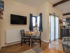 A room with a dining table, two chairs, a flat-screen TV mounted on the wall, a window with curtains, and a kitchen area with cabinets and a countertop. - Bugle (Saltburn-by-the-Sea)