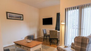 Ett vardagsrum med ett soffbord i trä, en grå soffa, ett matbord med två stolar, en tv som är monterad på väggen och ett fönster med gardiner.