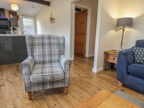 Una sala de estar con un sillón de cuadros, un sofá azul, piso de madera, una zona de cocina con un refrigerador y una puerta que da a otra habitación.