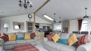 A cozy living room with two grey sofas, a red plaid ottoman, and a kitchen in the background.