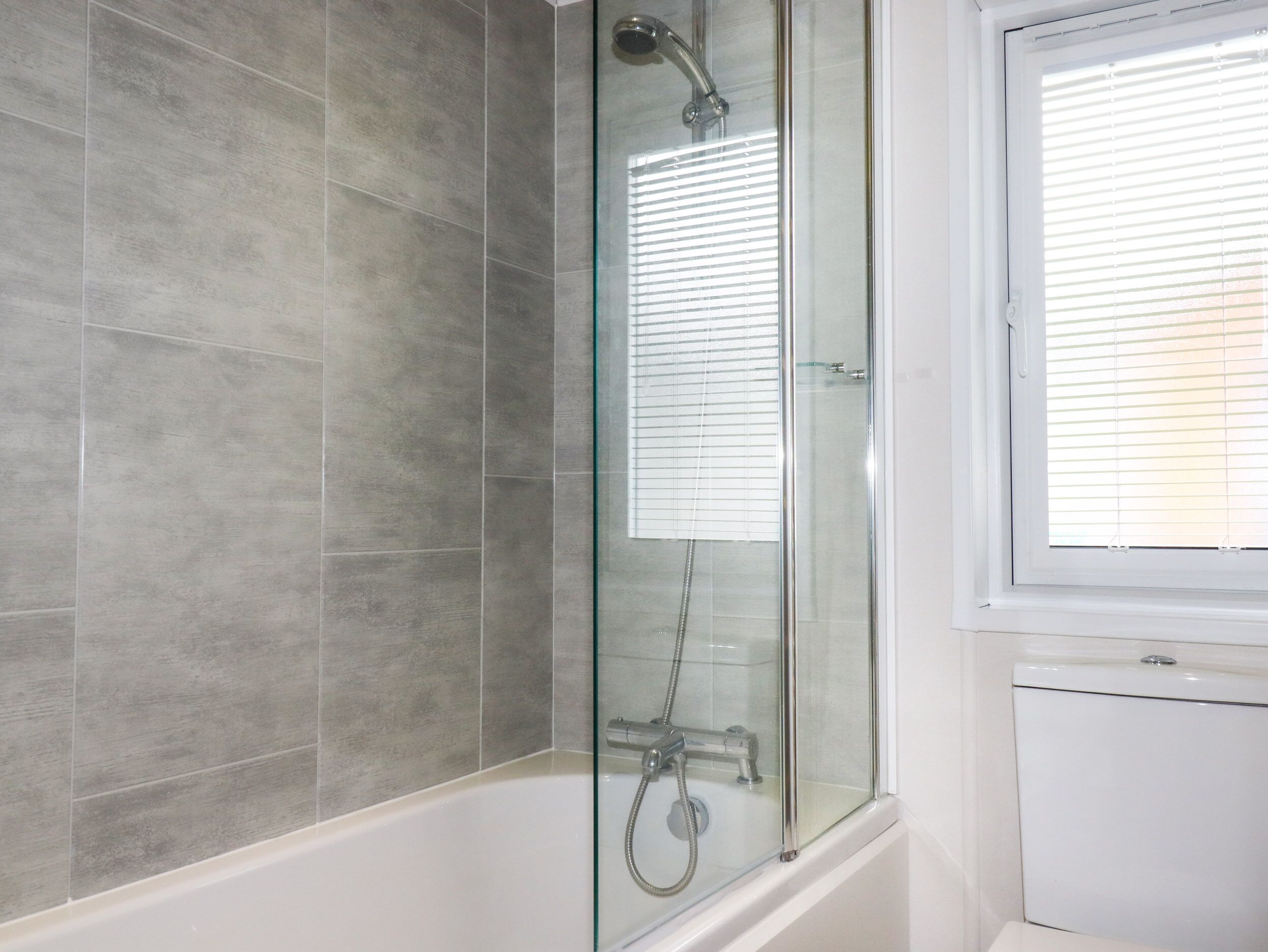 A modern bathroom with a glass-enclosed shower, grey tiles, and a window with blinds.