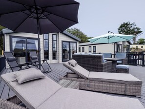 A covered outdoor seating area with lounge chairs and a table, surrounded by a deck and a building with large windows. - Beachcliff (Newquay)