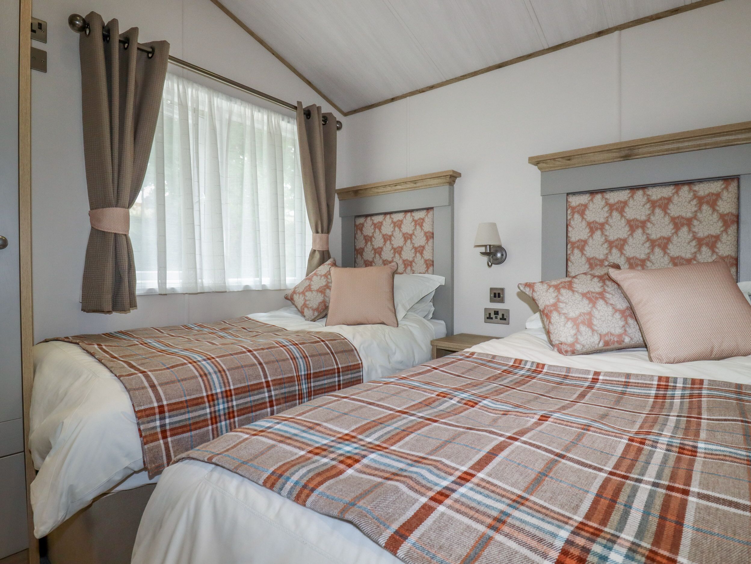 Une chambre double avec deux lits, chacun recouvert d’une couverture à carreaux, une table de chevet, une lampe fixée au mur et une fenêtre avec des rideaux.