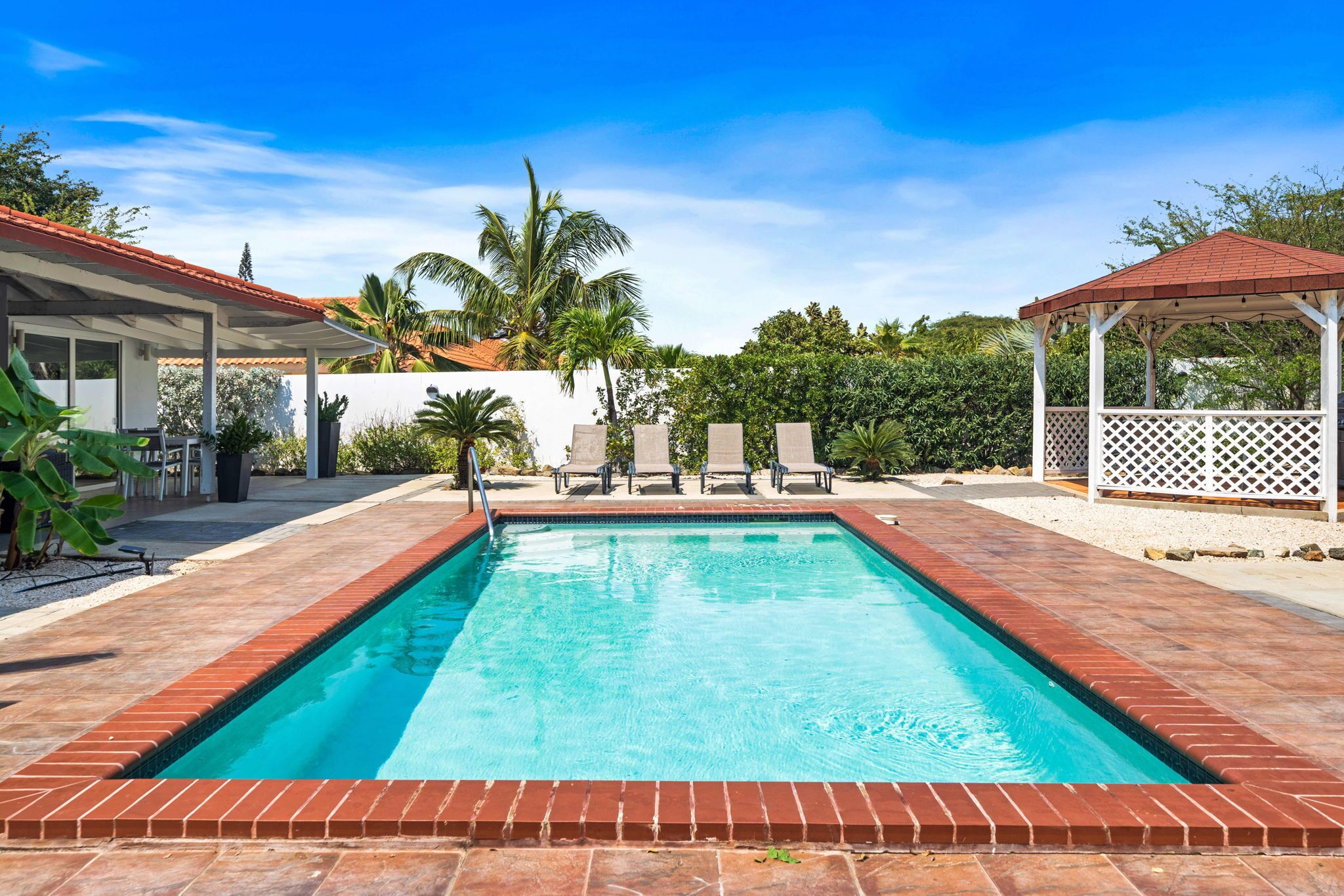 Una piscina rectangular con borde de azulejos, rodeada por un patio con sillas y un pabellón.