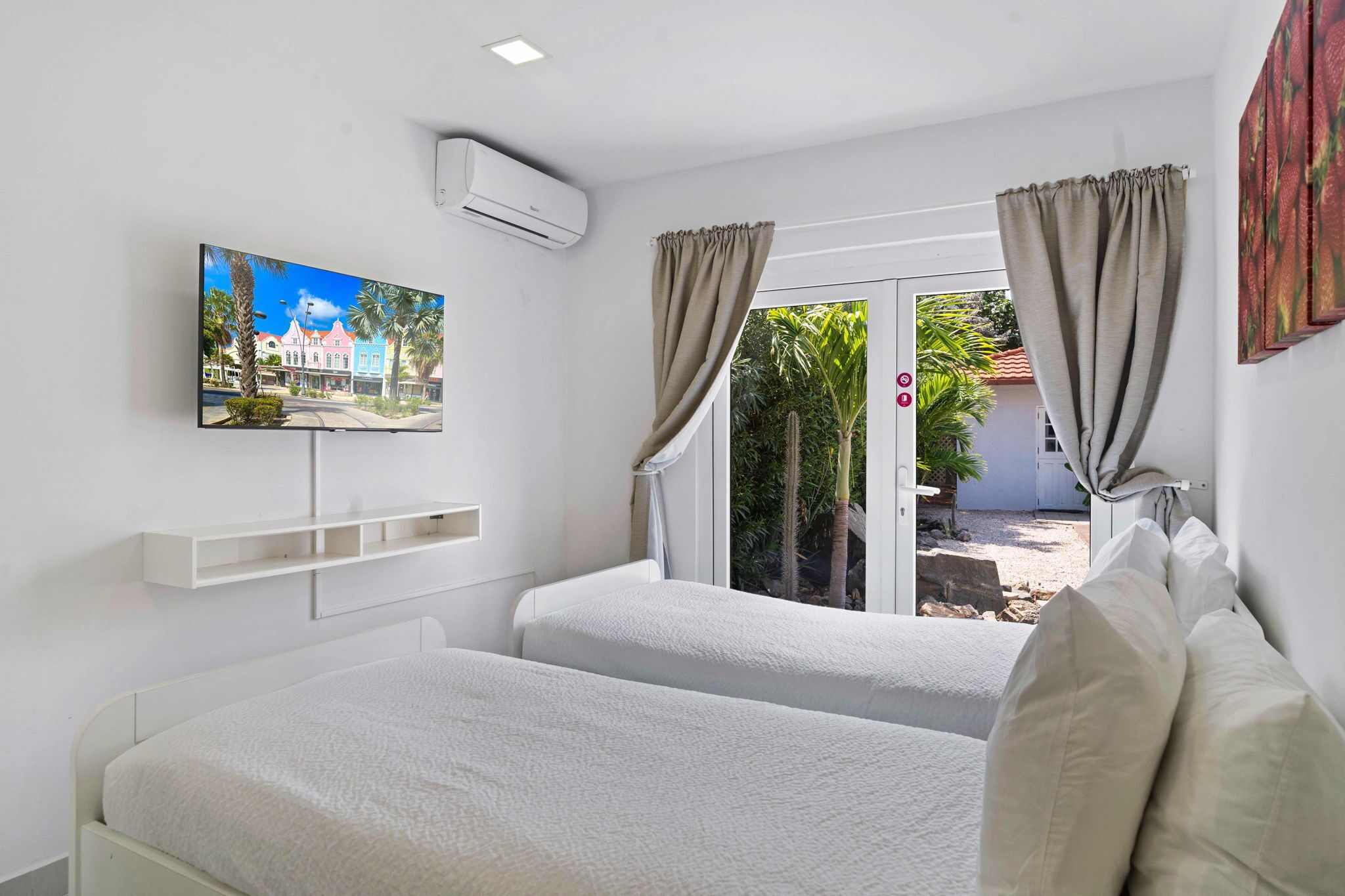 A hotel room with two beds, a TV on the wall, and a view of a garden through the door.