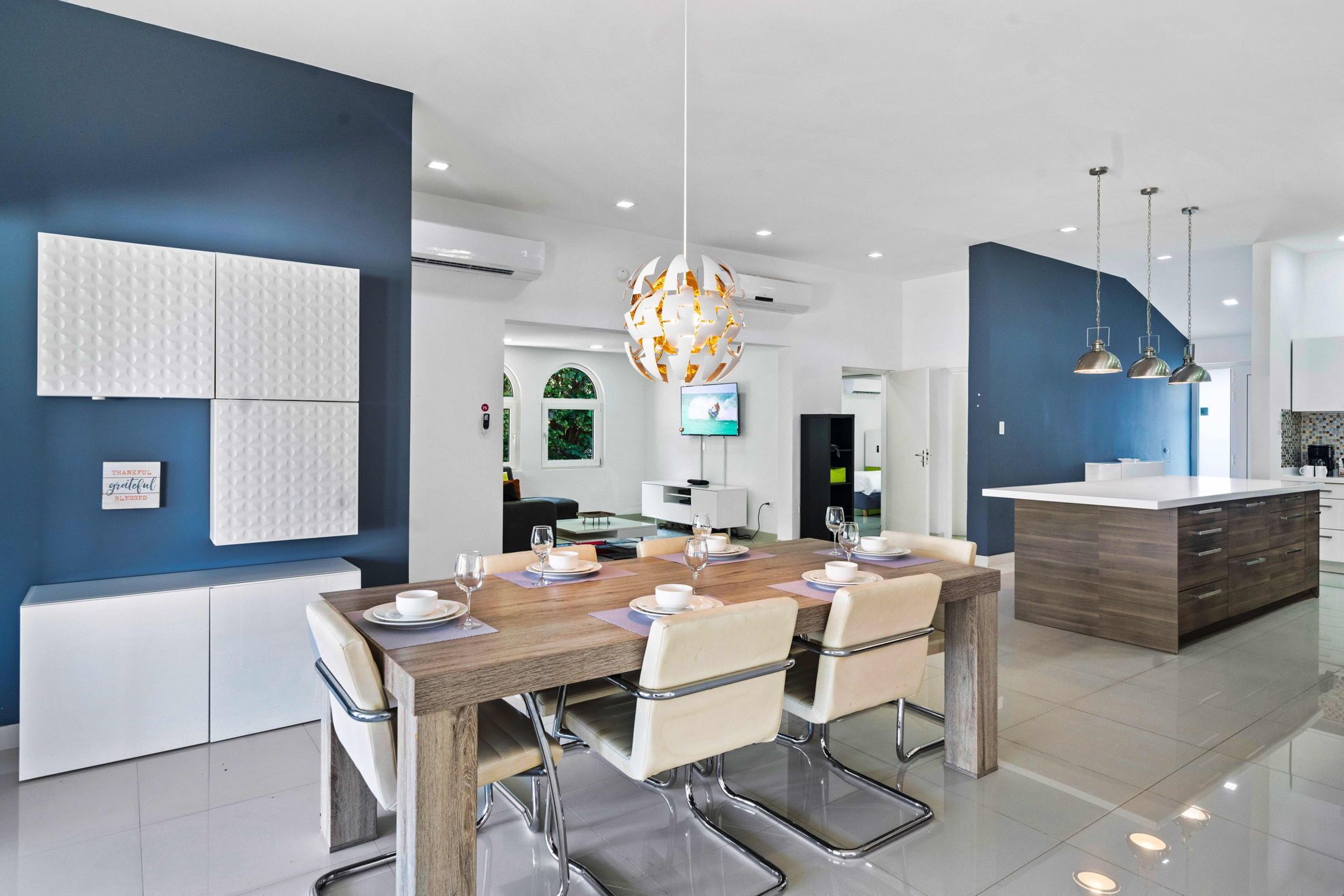 A modern dining area with a wooden table set for four, white cushioned chairs, a central chandelier, and a kitchen island in the background.