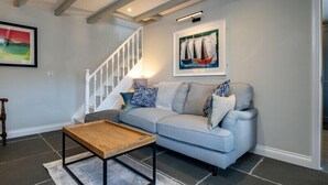 A living room with a light blue sofa, a wooden coffee table, a staircase, and a framed painting of sailboats. - Mino Cottage (Port Isaac)
