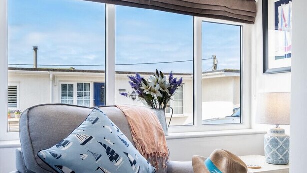 A cozy room with a grey armchair, a blue patterned pillow, a hat, and a vase with flowers. - Mino Cottage (Port Isaac)