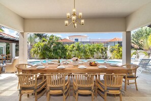 An outdoor dining area with a wooden table set for six, surrounded by wicker chairs, overlooking a swimming pool with palm trees and a building in the background.