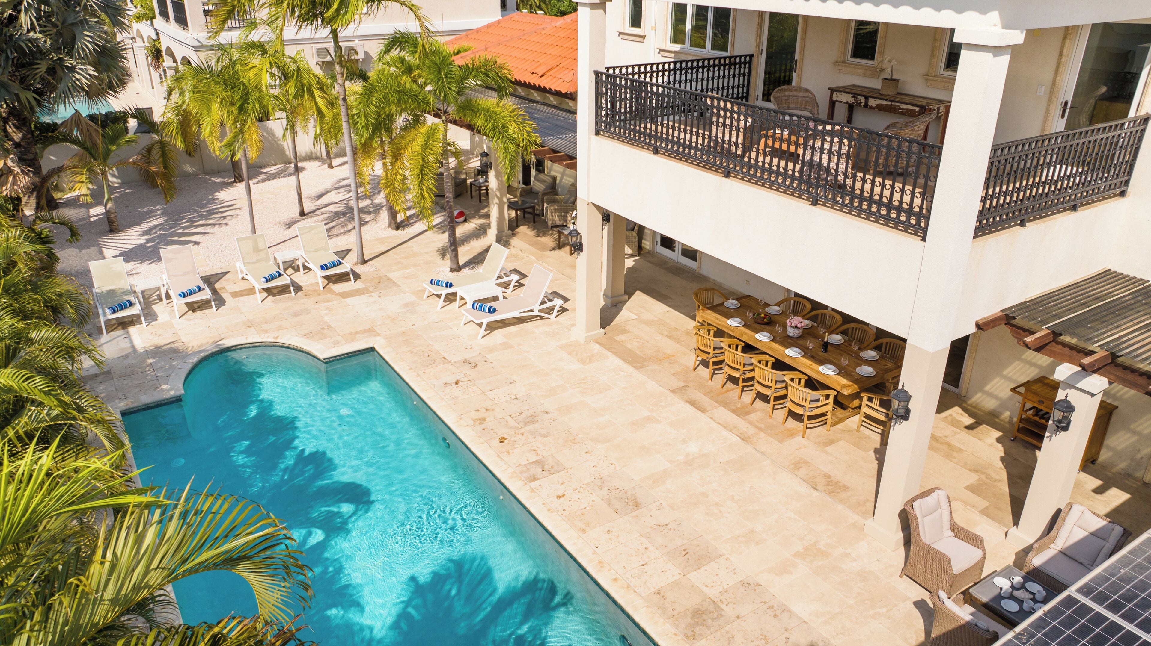 A pool area with lounge chairs and a dining table, surrounded by palm trees.