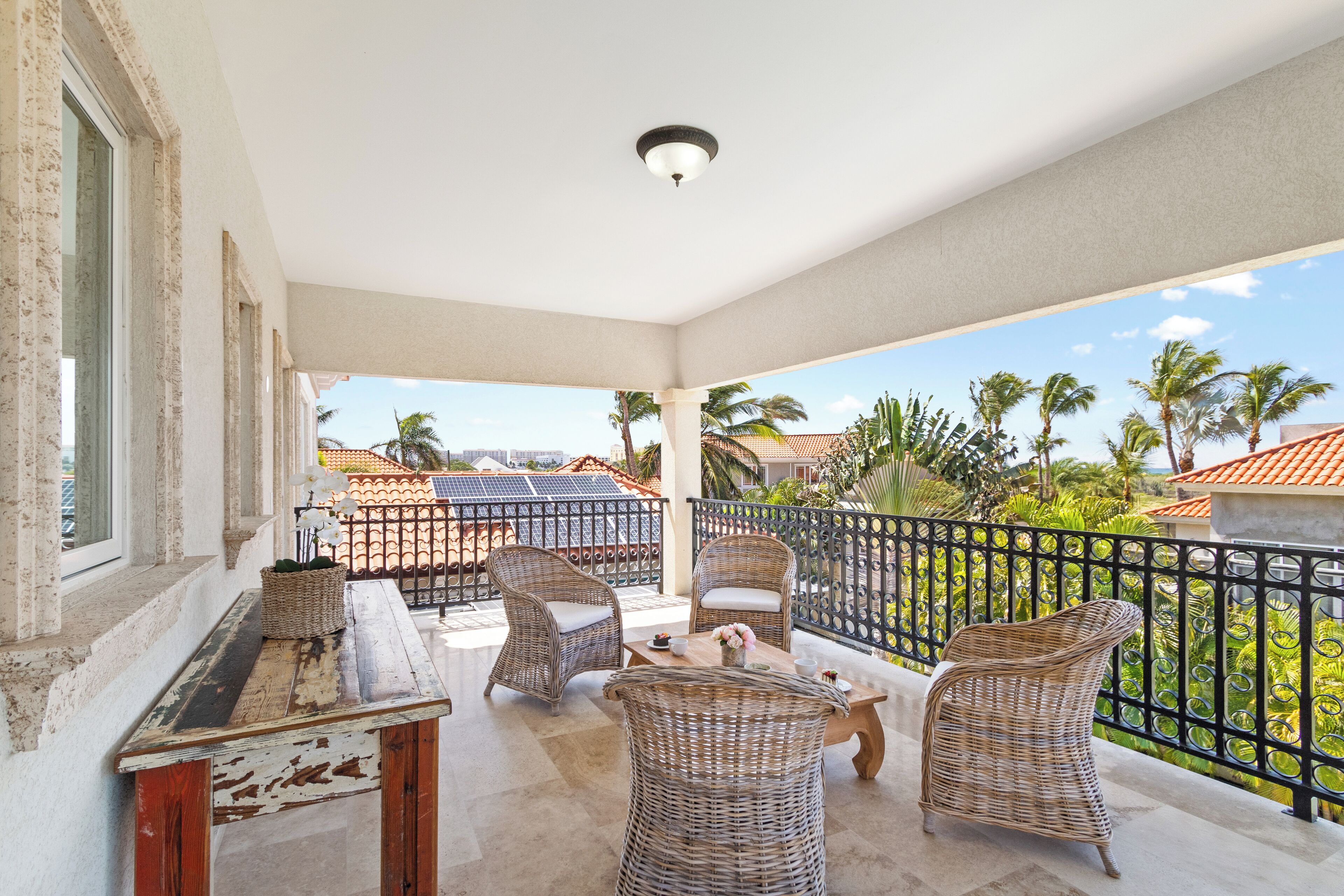 A balcony with wicker furniture and a table, overlooking a residential area with palm trees and houses.