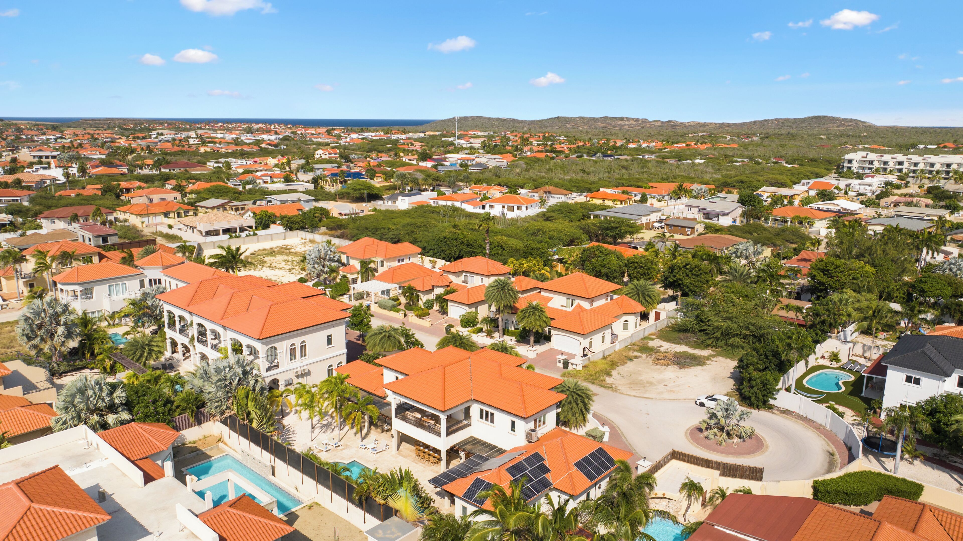 Un quartier résidentiel où les maisons se distinguent par leurs toits recouverts de tuiles rouges, le tout étant entouré de verdure et surplombé par un ciel dégagé.