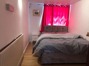 Une chambre à coucher avec un lit, une table de chevet, un radiateur, une fenêtre avec des rideaux rouges et un tableau encadré au mur.