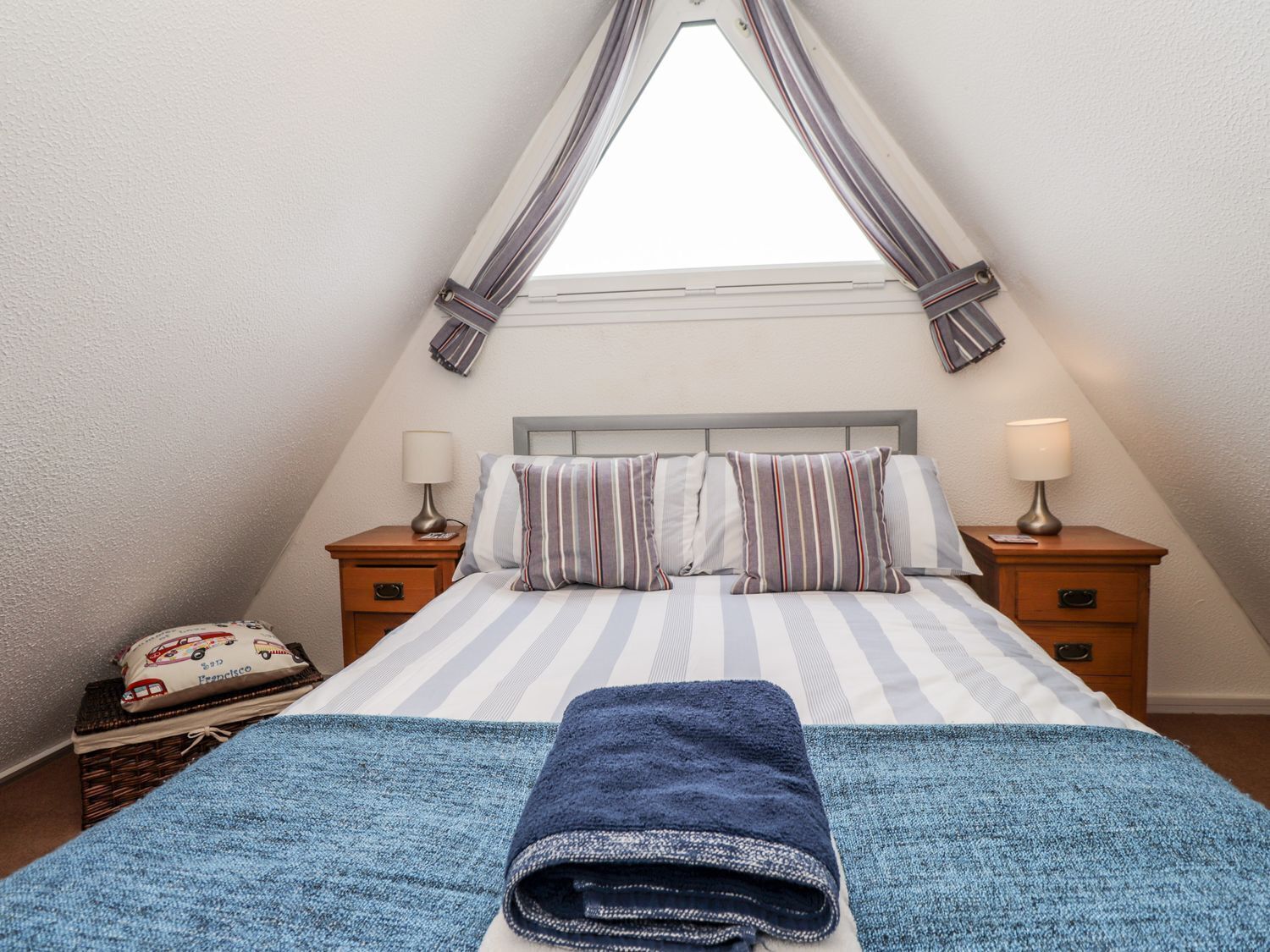 A bedroom with a bed, two bedside tables, a window with curtains, and a blue towel on the bed.