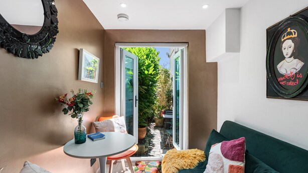 A modern living room with a green sofa, a small round table with a vase and a book, a mirror, and a door leading to a garden. - Tiny House, Brighton With Parking (Brighton)