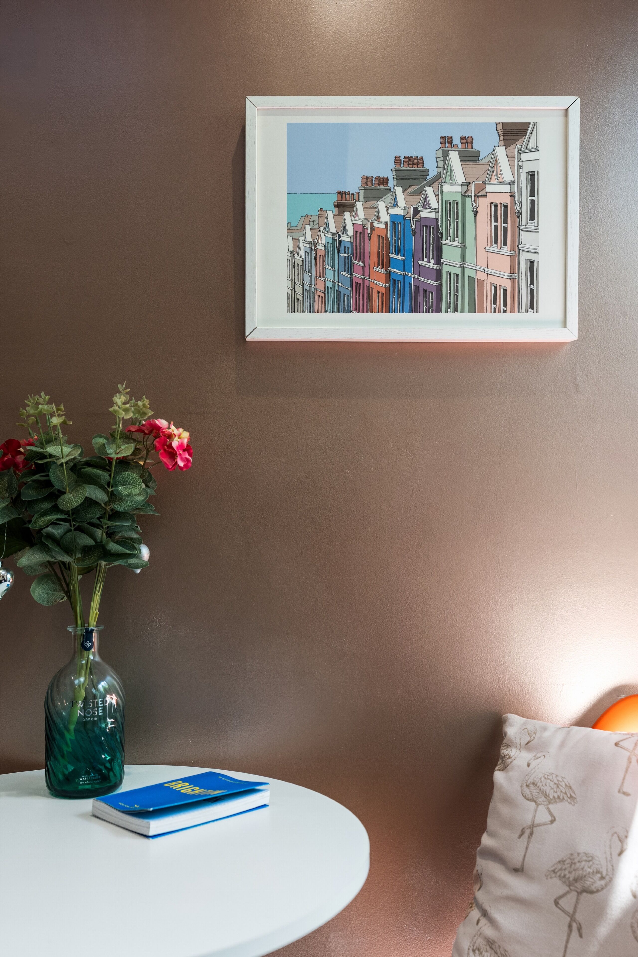 A framed artwork of colorful houses on a wall, a vase with flowers, a book, and a bottle on a table.
