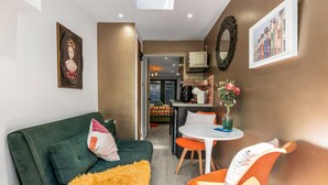 A compact living space with a green sofa, orange chairs, a small dining table, and a kitchen area in the background. - Tiny House, Brighton With Parking (Brighton)