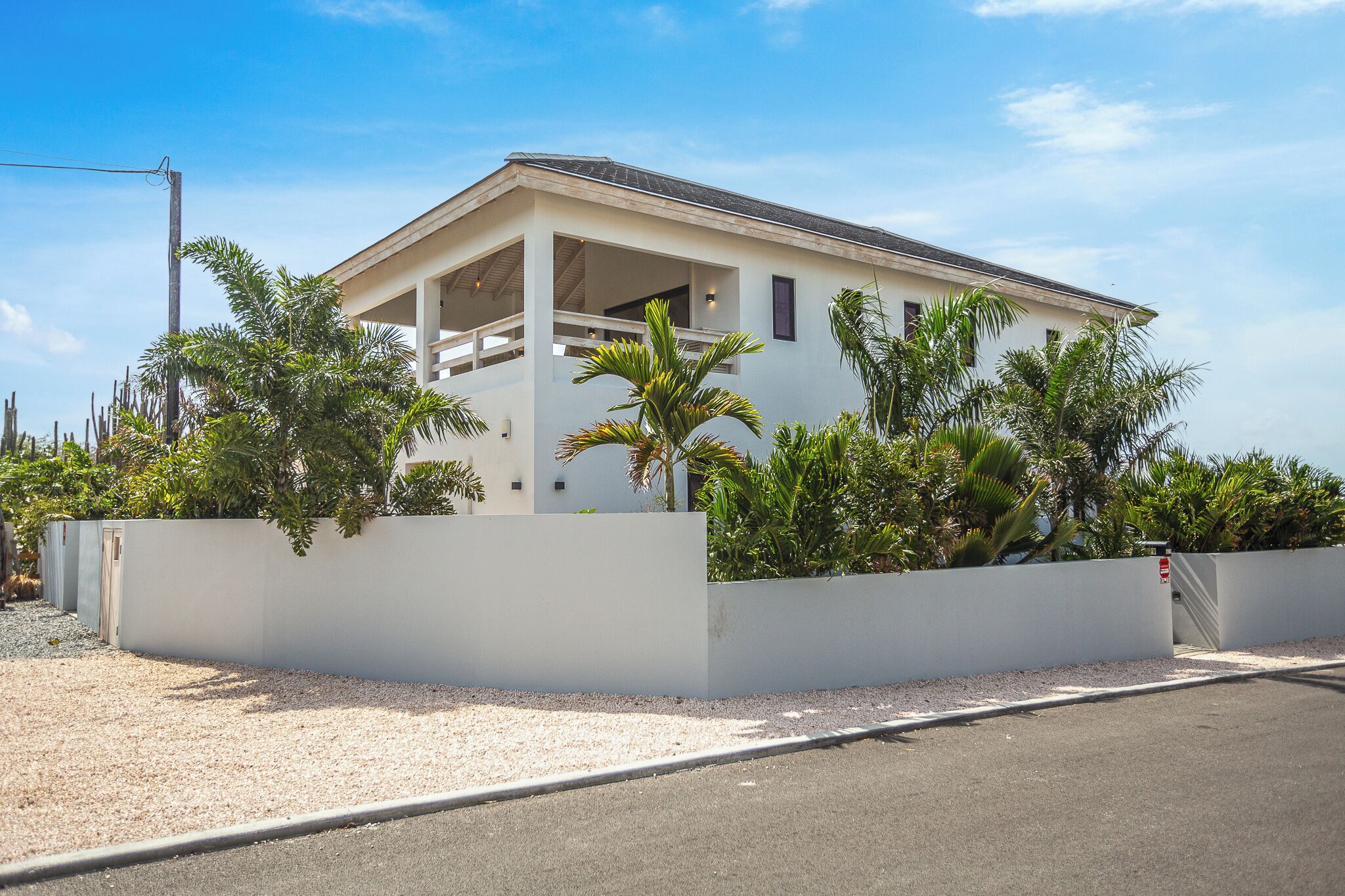 A modern two-story house with a balcony, surrounded by a white wall and tropical plants.