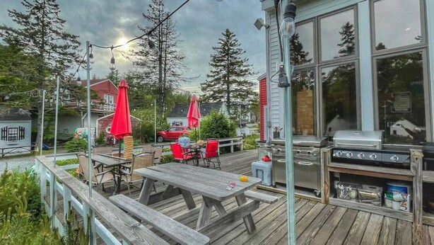 Terraza exterior con mesas de madera y sombrillas rojas, una parrilla de barbacoa y vistas a casas y árboles.