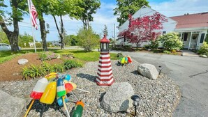 Un jardín con un faro rojo y blanco a rayas, boyas de colores y un edificio blanco con techo rojo.