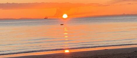 A beach scene with a sunset over the water, a boat, and a distant shoreline.