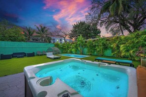 Un jacuzzi en un patio trasero con piscina, rodeado de vegetación y una valla verde.