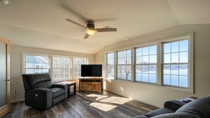 A spacious living room with a large window, a ceiling fan, and a flat-screen TV on a wooden stand. - Newly Built Lake Front Home (Lake Pleasant)