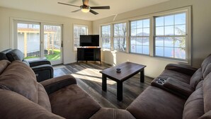 A living room with a ceiling fan, a flat-screen TV on a stand, a coffee table, and large windows with a view of a lake. - Newly Built Lake Front Home (Lake Pleasant)