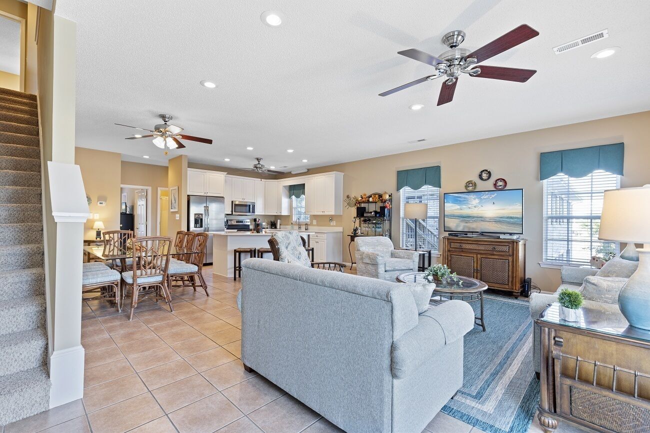 A spacious living room with a grey sofa, a dining area with wicker chairs, a kitchen with white cabinets, and a ceiling fan.