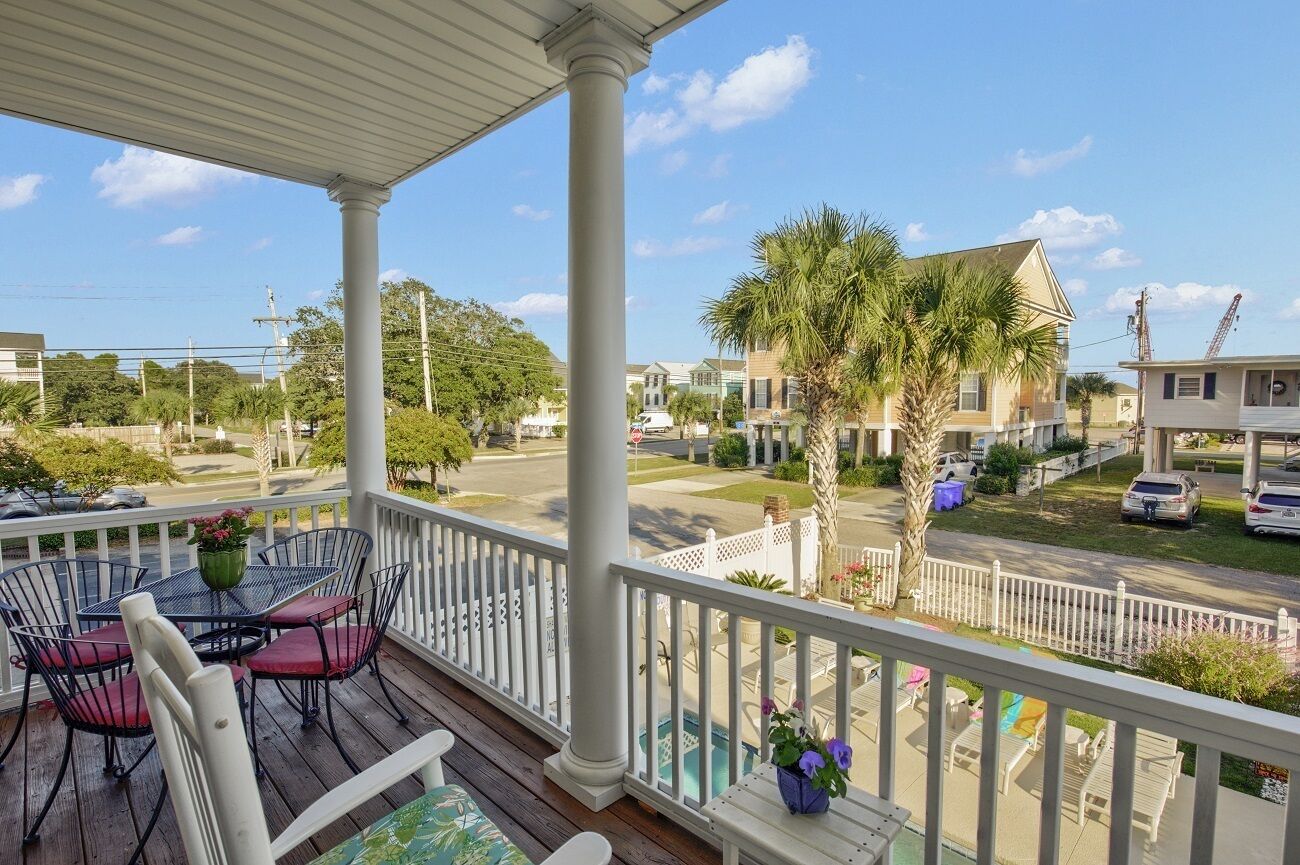 A covered porch with a table and chairs, overlooking a residential street with houses and palm trees.