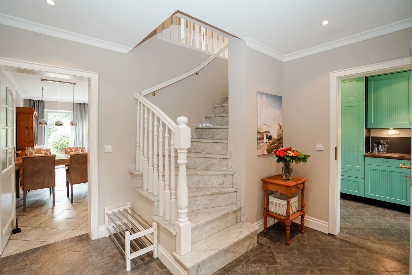 Una escalera con balaustres blancos, un comedor con mesa y sillas de madera, y una cocina con gabinetes color turquesa.