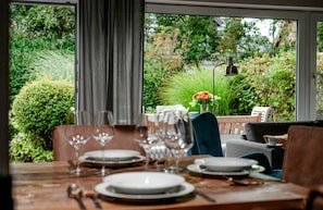 Uma sala de jantar com uma mesa posta para quatro pessoas, uma cadeira e vista para um jardim através de grandes janelas.