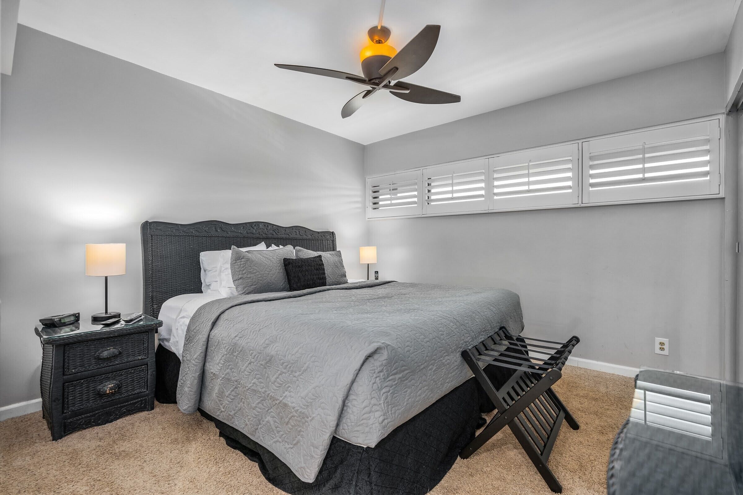 Un dormitorio con cama, mesitas de noche, ventilador de techo y ventana con persianas.