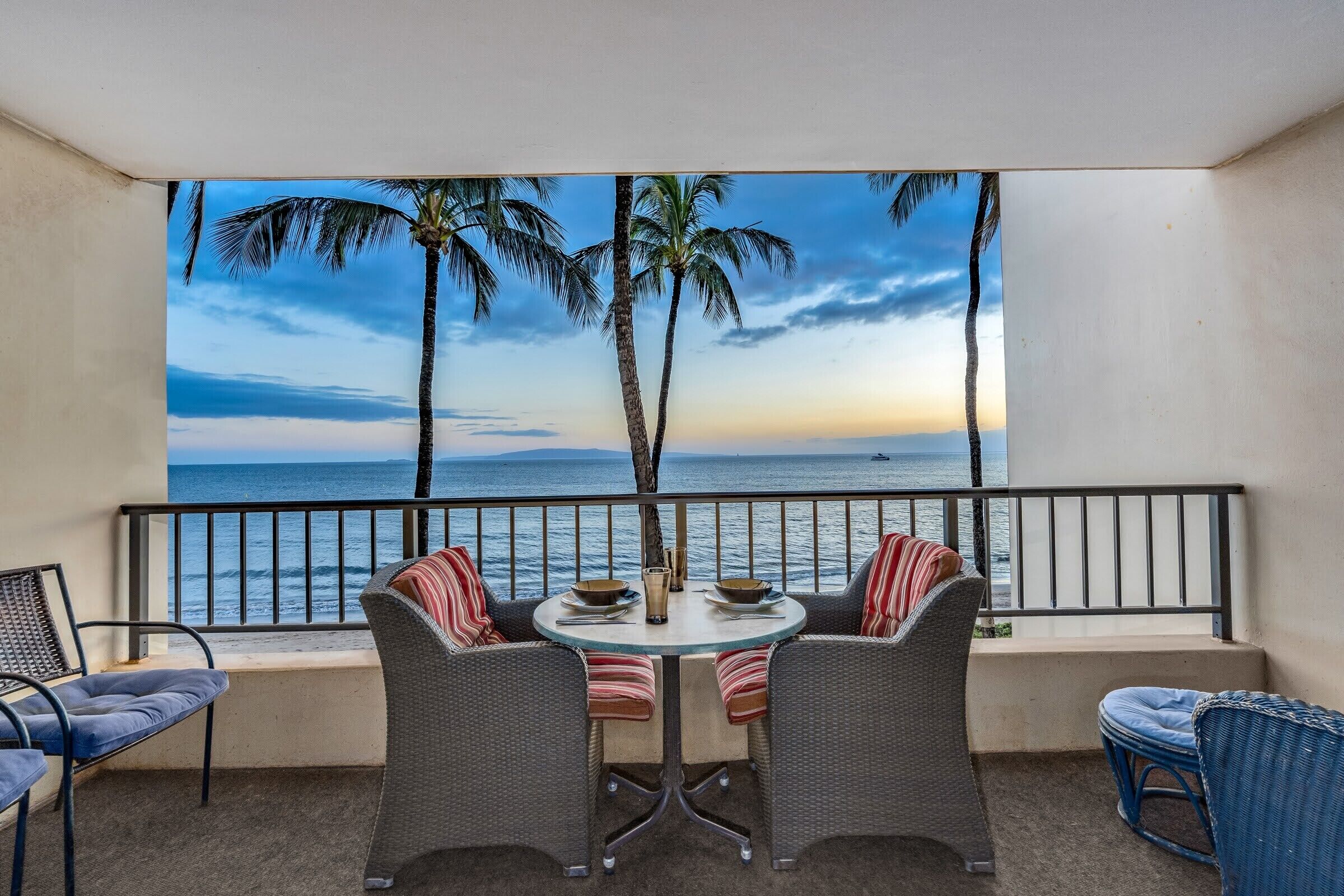 A balcony with wicker furniture, a round table, and a view of the ocean with palm trees.