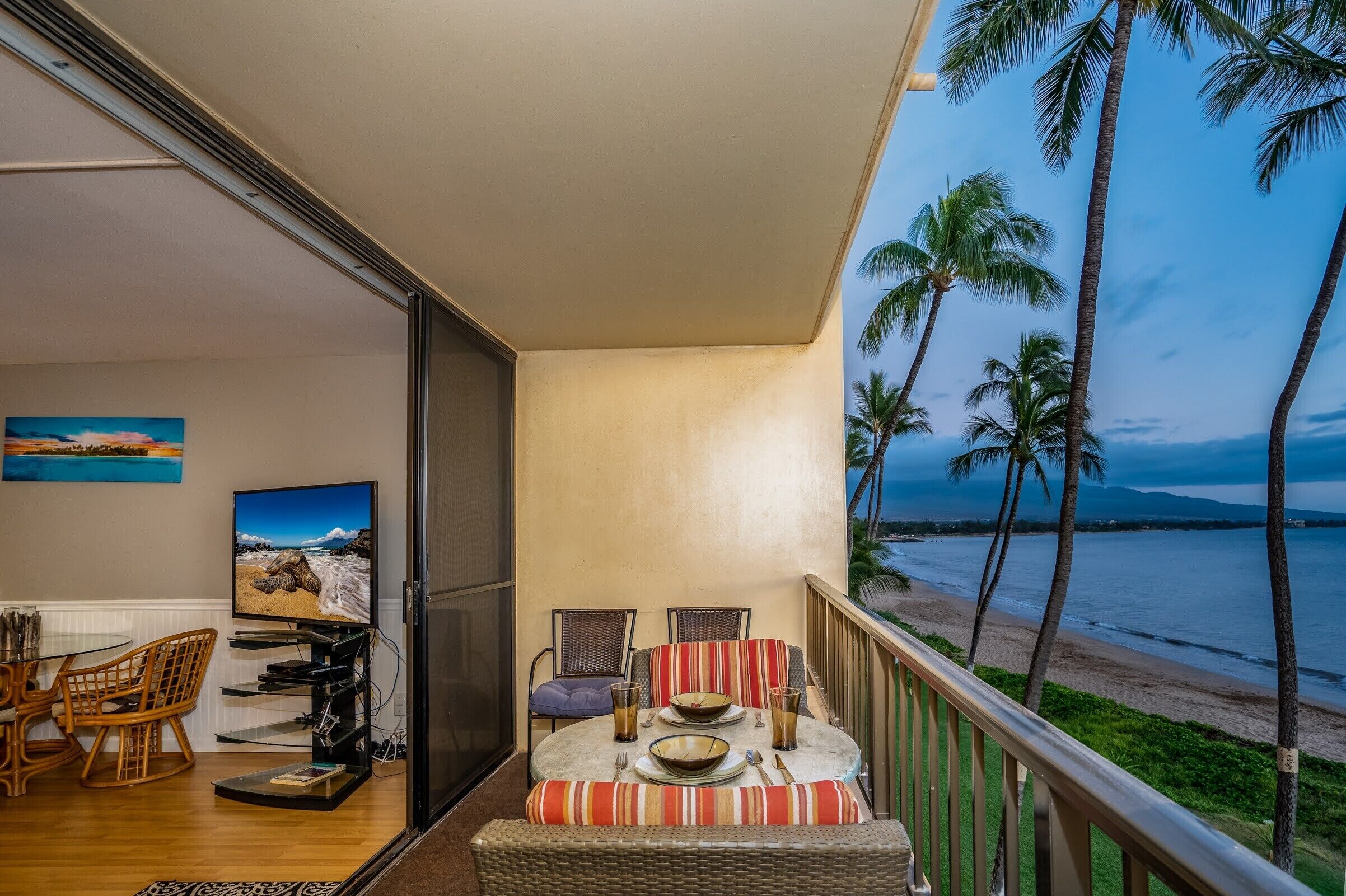 A balcony with a dining area, a television, and a view of the beach and palm trees.