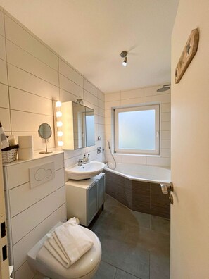 Une salle de bain moderne comprenant un meuble-lavabo, un lavabo et un miroir avec éclairage intégré, des toilettes et une baignoire placée sous une fenêtre.