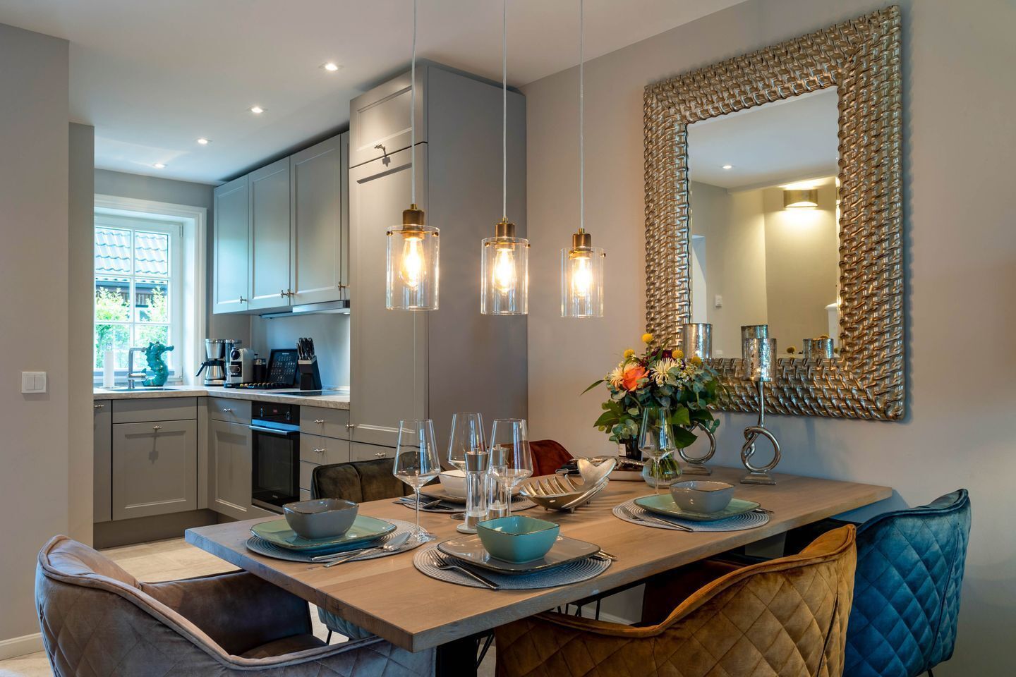Een moderne eetruimte met een houten tafel voor vier personen, een keuken op de achtergrond en hanglampen erboven.