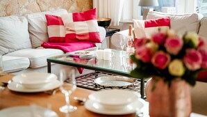 A well-lit living room with a glass coffee table, white sofas, and red and white cushions. A vase of flowers is on the coffee table, and there are plates, glasses, and a bottle on the table. - Haus Sprotte - Welcome to the "Sprotte" house (Sylt-Ost)