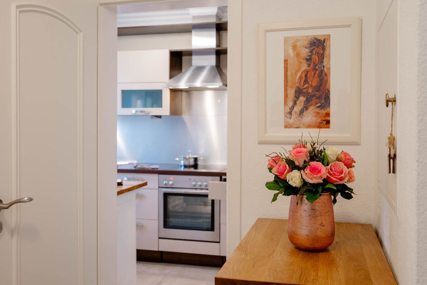 A kitchen with stainless steel appliances, a wooden table with a vase of flowers, and a framed painting on the wall.