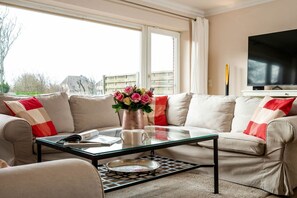 En stue med en beige sofa, et glasbord og et fladskærms-tv på et stativ.