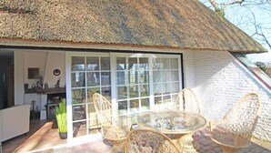 Une maison à toit de chaume avec une porte vitrée donnant sur un patio aménagé avec des meubles en osier et un pot de fleurs.