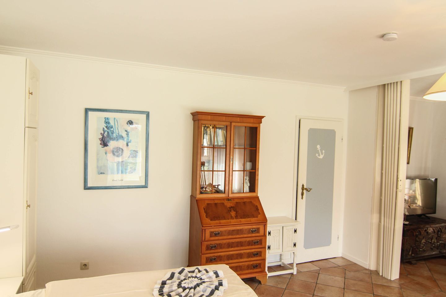 A bedroom with a bed, a wooden cabinet, a framed picture, a door with an anchor symbol, and a television in the corner.
