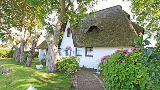 Une maison de campagne traditionnelle, aux murs blancs et au toit de chaume, entourée d’une végétation luxuriante et d’arbres.