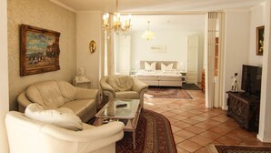 Un salon avec un canapé, des fauteuils, une table basse et un téléviseur. On aperçoit, au fond, une chambre à coucher avec un lit et un lustre.