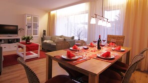 Un comedor con una mesa de madera dispuesta para cuatro personas, un sofá, un televisor y una ventana con cortinas.